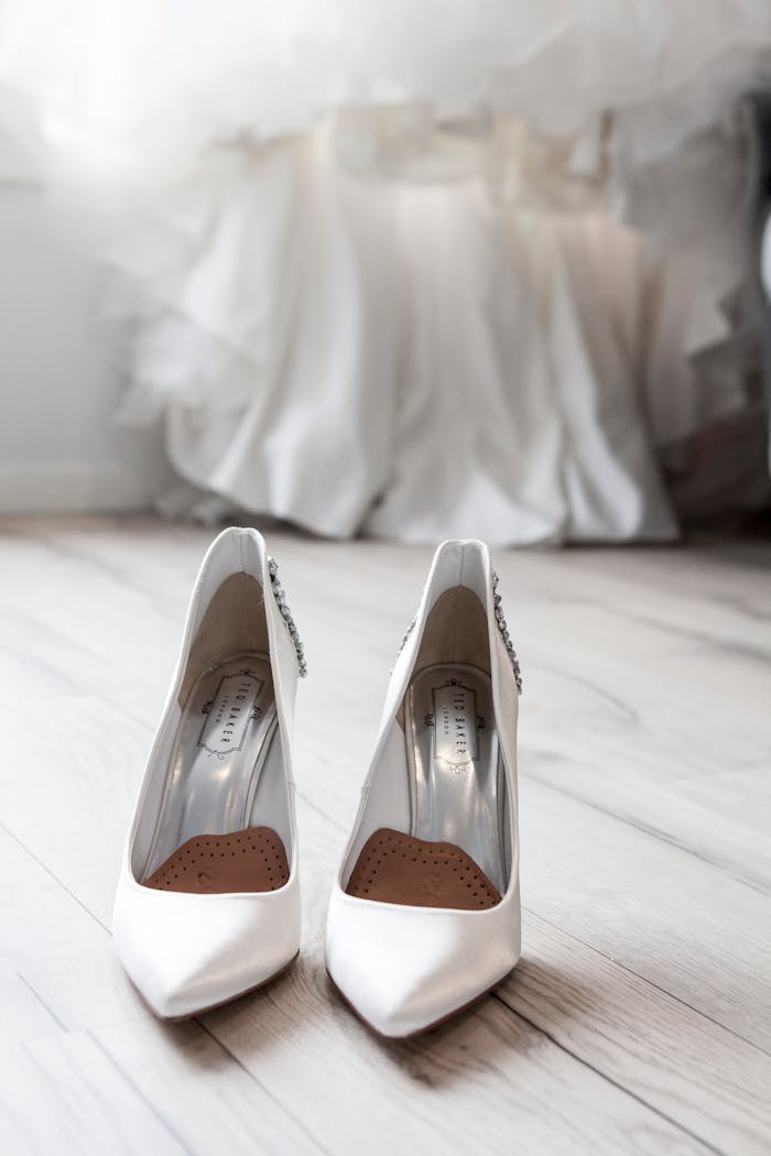 Stylish white bridal shoes positioned in front of a flowing wedding dress indoors.